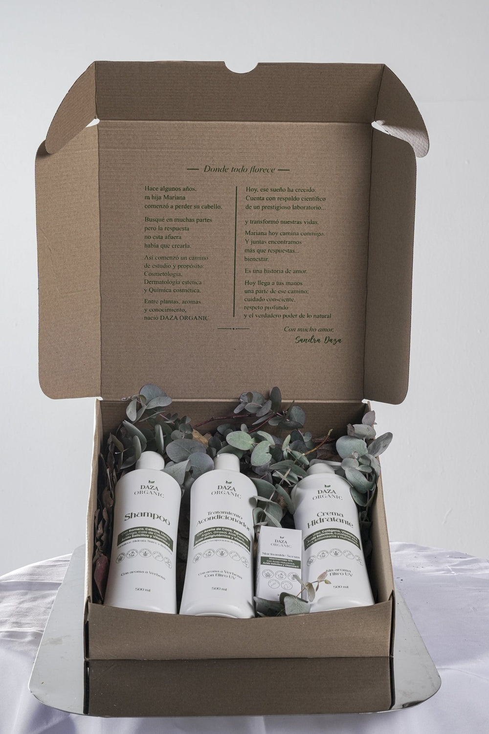 Caja de cartón abierta con los productos de Daza Organic entre hojas de eucalipto; en la tapa, la carta impresa «Donde todo florece».