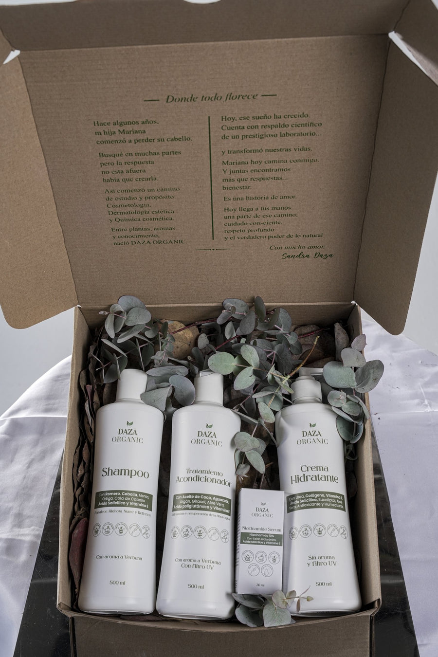 Caja kraft de Daza Organic abierta, con la carta «Donde todo florece» en la tapa; dentro, Shampoo Nutritivo, Tratamiento Acondicionador, la caja del Niacinamide Serum y Crema Hidratante entre ramas de eucalipto.