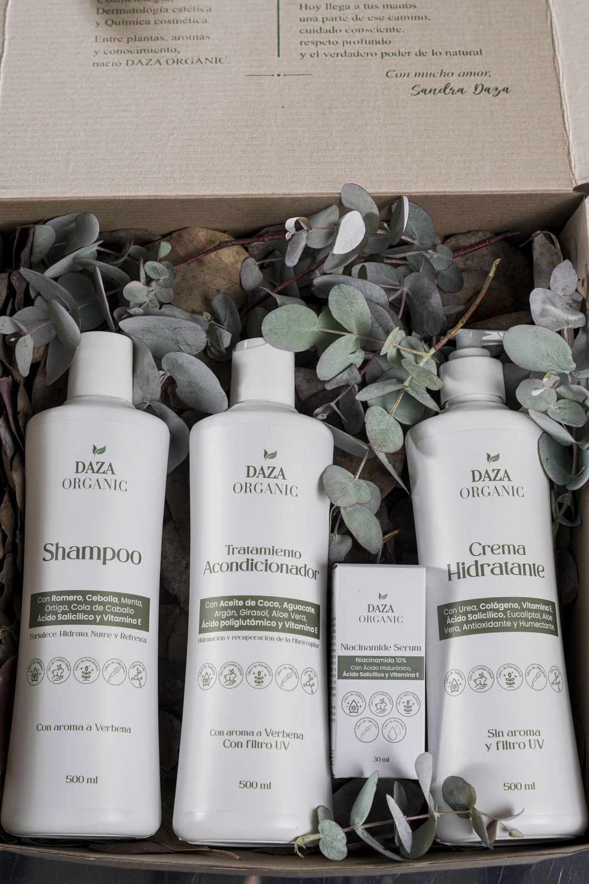 Primer plano de una caja de cartón con el shampoo, el tratamiento acondicionador, la caja del Niacinamide Serum y la crema hidratante de Daza Organic entre hojas de eucalipto.