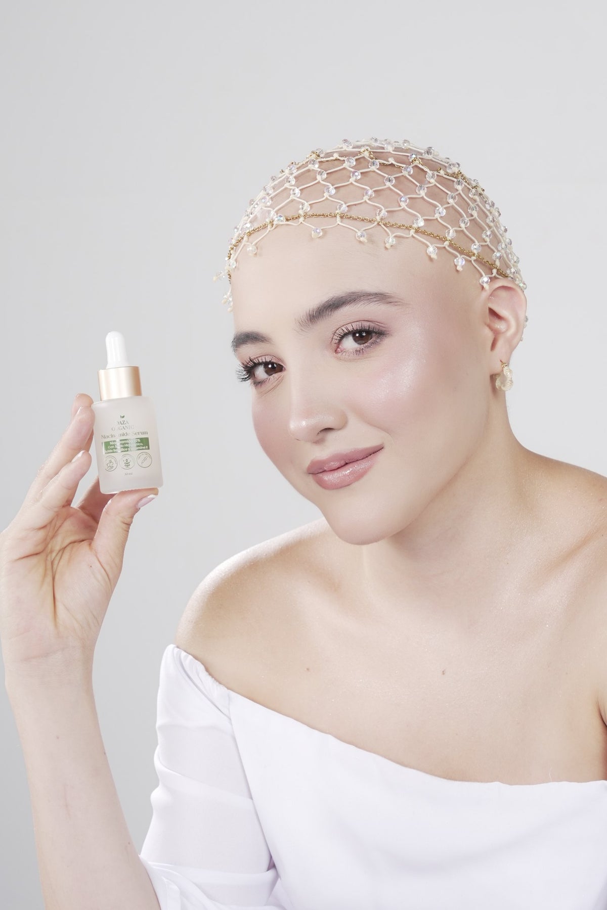 Mujer con un tocado de red de cuentas sonríe e inclina la cabeza mientras muestra el frasco gotero del Niacinamide Serum de Daza Organic.