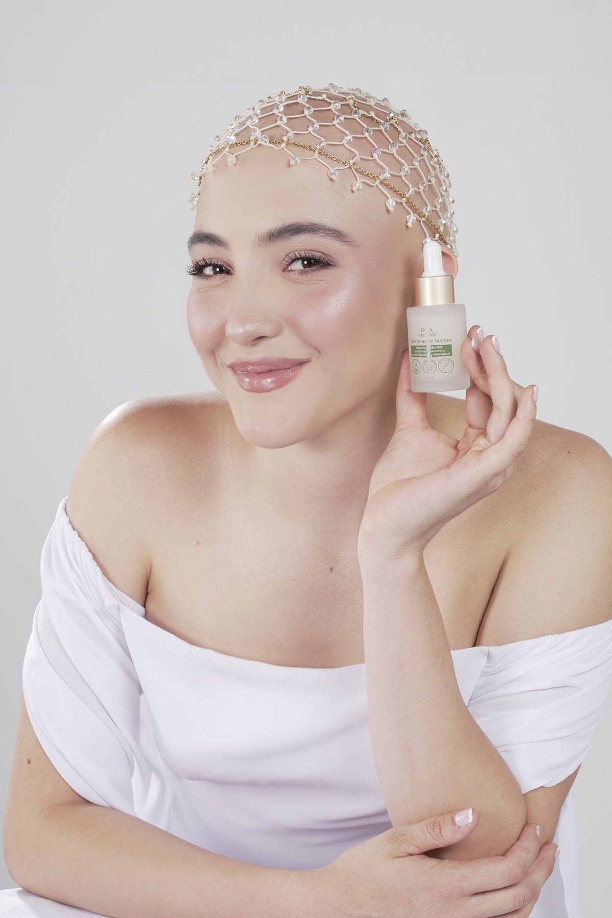Mujer con un tocado de red de cuentas sonríe y sostiene el Niacinamide Serum de Daza Organic junto a la mejilla; viste un top blanco de hombros descubiertos sobre fondo gris claro.