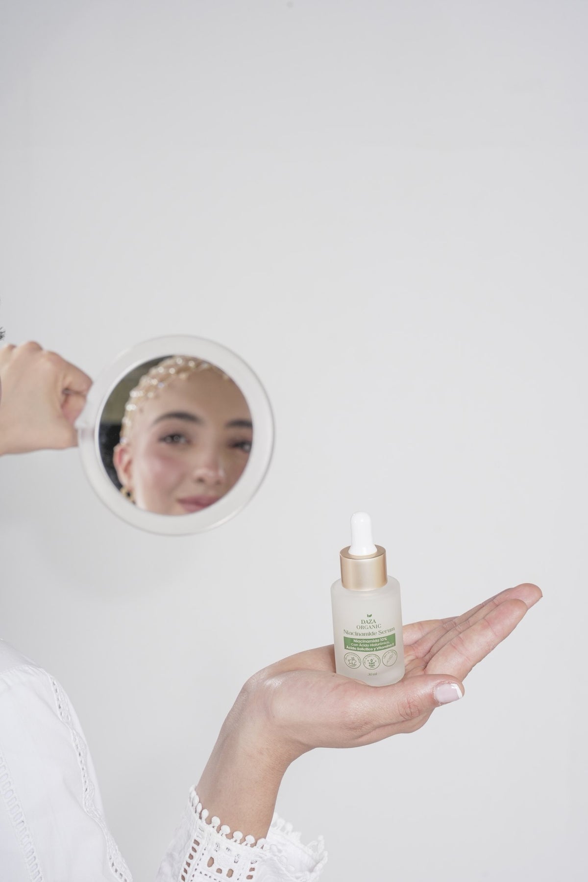 Una mano sostiene en la palma el frasco gotero del Niacinamide Serum de Daza Organic; detrás, otra mano levanta un espejo redondo que refleja el rostro de la modelo.