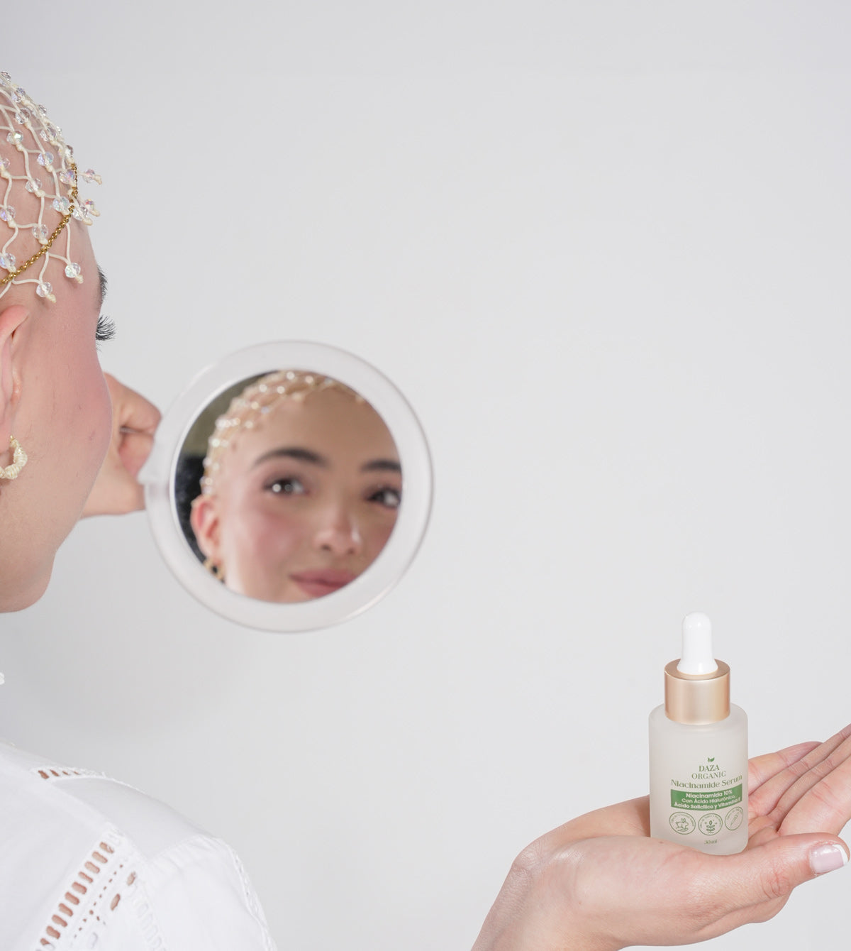 Una joven con una redecilla de cuentas sobre la cabeza se mira en un espejo de mano redondo; en primer plano, una mano sostiene en la palma el frasco del Niacinamide Serum de Daza Organic.